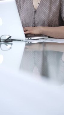 Kas yığını noktalı bluz giyen iş kadını, dizüstü bilgisayarda daktilo kullanıyor, vergileri ve denetim raporlarını hazırlıyor, cam ofis masasında dokümanlar, dikey görüş. Vergiler, iş denetimleri.