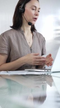 Kaslı noktalı bluz giyen iş kadını, modern ofis ortamında, dikey portrede çalışırken bilgisayarına odaklanmış bir kulaklık takıyor. İş adamları konsepti.