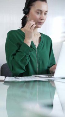 Yeşil bluzlu iş kadını, modern ofis ortamında, dikey portrede çalışırken bilgisayarına odaklanmış bir kulaklık takıyor. İş adamları, çağrı merkezi kavramı.