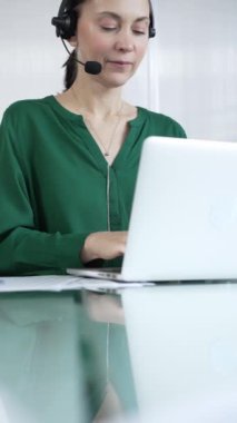 Yeşil bluzlu iş kadını, modern ofis ortamında, dikey portrede dizüstü bilgisayarla etkin bir şekilde çalışırken, bir kulaklık takıyor. İş adamları, çağrı merkezi kavramı