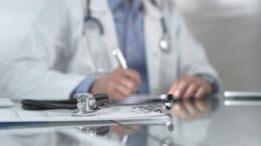 Stetoskop ve ilaç geçmişi kayıtları cam bir masanın üzerinde duruyor. Klinik ofis arka planında dizüstü bilgisayarla çalışan bir erkek doktorun önünde. Tıp konsepti.