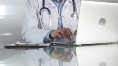 Doktor kadın, doktor önlüğünün altına mavi bir bluz giyiyor, sağlık hizmetlerinde teknolojiyi kullanıyor, elleri bilgisayarında stetoskop, gözlük ve pano ile daktilo kullanıyor. Tıp konsepti.