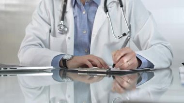 Beyaz bir sağlık önlüğünün altına mavi gömlek giyen erkek doktor, bir not panosu kullanıyor ve yakın plandaki cam masaya notlar alıyor. Tıp konsepti.
