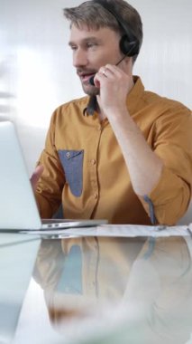 Sarı gömlekli bir iş adamı, modern ofis ortamında dikey portre üzerinde çalışırken bir dizüstü bilgisayara odaklanmış bir kulaklık takıyor. İş adamları konsepti.