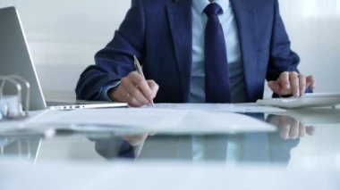 İş adamı hesap makinesi kullanıyor ve not alıyor. Ofis masasında dizüstü bilgisayar ve belgelerle çalışıyor. İş dünyasında denetim ve vergi teması.