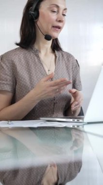 Kaslı noktalı bluz giyen iş kadını, modern ofis ortamında, dikey portrede çalışırken bilgisayarına odaklanmış bir kulaklık takıyor. İş adamları konsepti.