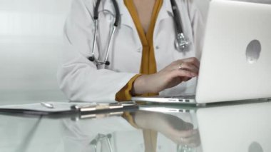 Tıbbi önlüğün altına sarı bluz giyen kadın doktor, sağlık hizmetlerinde teknolojiyi kullanıyor, bilgisayarında stetoskop, gözlük ve not panosuyla yazı yazan elleri kapalı. Tıp konsepti.