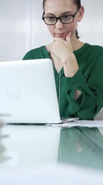Yeşil bluzlu odaklanmış iş kadını dizüstü bilgisayar kullanıyor ve çene üzerinde elle düşünüyor, parlak bir ofis, dikey portre üzerinde çalışıyor. İş adamları konsepti.