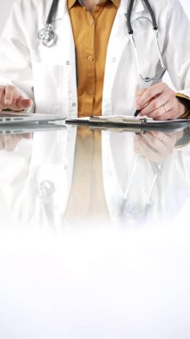 Stetoskop giyen, laboratuvar önlüğünün altına sarı tişört giyen kimliği belirsiz bir erkek doktor, pano kullanıyor ve tıp bürosundaki cam masaya notlar alıyor. Tıp konsepti.