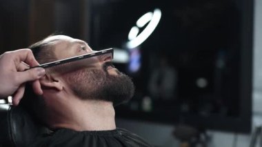 Profesyonel berber, modern berber dükkanında yüksek kaliteli tımar hizmetleri sunarak gevşetilmiş müşterinin sakalını elektrikli jiletle dikkatlice buduyor.