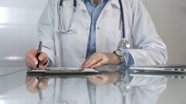 Beyaz bir sağlık önlüğünün altına mavi bir bluz giyen kadın doktor, bir pano kullanıyor ve yakın plandaki cam masaya notlar alıyor. İlaç..