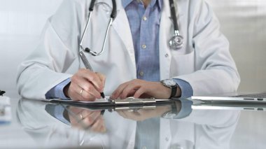 Beyaz bir sağlık önlüğünün altına mavi gömlek giyen erkek doktor, bir not panosu kullanıyor ve yakın plandaki cam masaya notlar alıyor. Tıp konsepti.