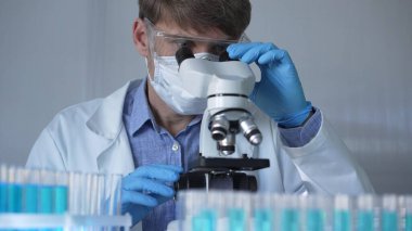 Laboratuvar önlüğü, maske, mavi eldiven ve güvenlik gözlüğü takan bir bilim adamı laboratuarda test tüpleriyle çevrili bir dürbün kullanıyor. Portre görüntüsü. Tıp ve Bilim.