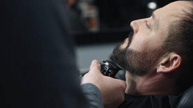 Profesyonel berber, modern berber dükkanında yüksek kaliteli tımar hizmetleri sunarak gevşetilmiş müşterinin sakalını elektrikli jiletle dikkatlice buduyor.