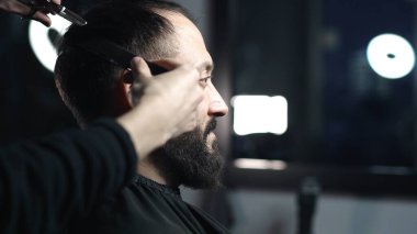 Profesyonel berber, modaya uygun bir berber dükkanında profesyonel bir makasla, sakallı bir adam için modern bir saç stili hazırlayan bir müşterinin saçını ustaca kesiyor.