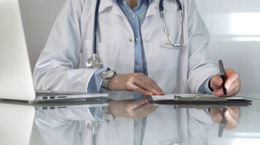 Beyaz bir sağlık önlüğünün altına mavi bir bluz giyen kadın doktor, bir pano kullanıyor ve yakın plandaki cam masaya notlar alıyor. İlaç..