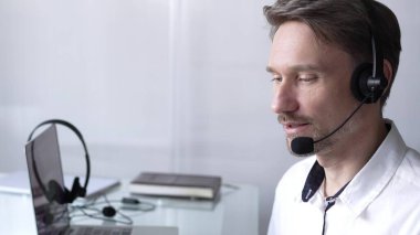 Kulaklık ve gözlük takan bir iş adamı, video konferans görüşmesi için laptop kullanıyor, merkez ofisten uzaktan çalışıyor. Çağrı merkezi kavramı.