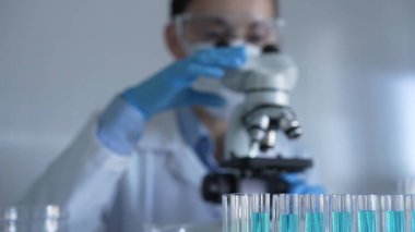 Mavi bir sıvıyla dolu laboratuvar tüpleri odaklanmış, yakından bakıyor. Kadın araştırmacı, arka planda mikroskop kullanıyor. Tıp ve bilim konsepti.