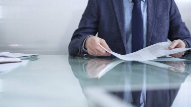 Mavi ceketli bir işadamı modern ofis ortamında cam bir masada belgeleri tutarken önemli evrakları inceliyor. İş adamları veya denetim kavramı.