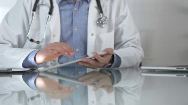 Tıbbi beyaz önlüğün altına mavi gömlek giyen doktor, modern sağlık ortamında dijital tablet kullanıyor, hasta verileriyle etkileşimde, yakın plan. Tıp konsepti.