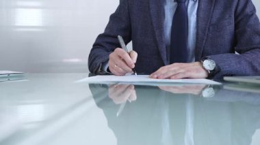 Çağdaş bir ofis ortamında modern cam bir masanın üzerinde profesyonel giyimli bir işadamı kalemle kontrat imzalıyor. İş adamları veya denetim kavramı