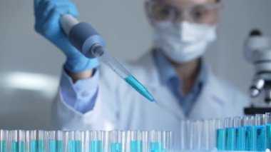 Laboratuvar önlüğü, maskesi ve güvenlik gözlüğü takan kadın araştırmacı test tüpüne mavi bir sıvı atmak için mikropipet kullanıyor. Tıp ve bilim konsepti.
