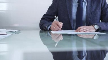 Çağdaş bir ofis ortamında modern cam bir masanın üzerinde profesyonel giyimli bir işadamı kalemle kontrat imzalıyor. İş adamları veya denetim kavramı