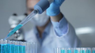 Laboratuvar önlüğü, maskesi ve güvenlik gözlüğü takan kadın araştırmacı test tüpüne mavi bir sıvı atmak için mikropipet kullanıyor. Tıp ve bilim konsepti.