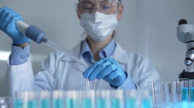 Laboratuvar önlüğü, maske ve güvenlik gözlüğü takan bilim adamı kadın araştırmacı test tüplerine sıvı atmak için bir mikropipet kullanıyor. Tıp ve bilim konsepti.
