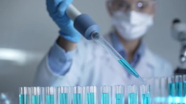 Laboratuvar önlüğü, maskesi ve güvenlik gözlüğü takan kadın araştırmacı test tüpüne mavi bir sıvı atmak için mikropipet kullanıyor. Tıp ve bilim konsepti.