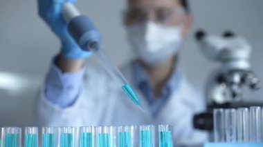 Laboratuvar önlüğü, maskesi ve güvenlik gözlüğü takan kadın araştırmacı test tüpüne mavi bir sıvı atmak için mikropipet kullanıyor. Tıp ve bilim konsepti.