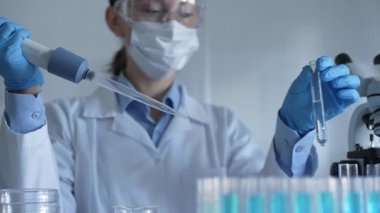 Laboratuvar önlüğü, maske ve güvenlik gözlüğü takan bilim adamı kadın araştırmacı test tüplerine sıvı atmak için bir mikropipet kullanıyor. Tıp ve bilim konsepti.