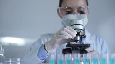 Laboratuvar önlüğü, maskesi, beyaz eldivenleri ve güvenlik gözlüğü takan bilim adamı bir kadın laboratuarda test tüpleriyle çevrili bir mikroskop kullanıyor. Portre görüntüsü. Tıp ve bilim konsepti.