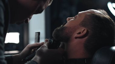 Profesyonel berber, modern berber dükkanında yüksek kaliteli tımar hizmetleri sunarak gevşetilmiş müşterinin sakalını elektrikli jiletle dikkatlice buduyor.