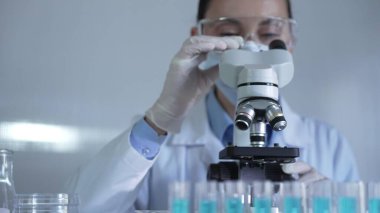 Laboratuvar önlüğü, maskesi, beyaz eldivenleri ve güvenlik gözlüğü takan bir bilim adamı kadın laboratuardaki test tüpleriyle çevrili bir mikroskobu ayarlıyor..