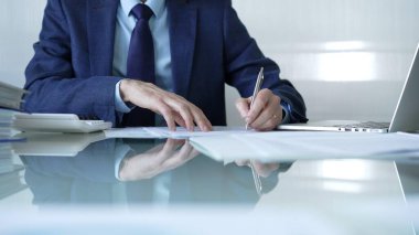 İş adamı hesap makinesi kullanıyor ve not alıyor. Ofis masasında dizüstü bilgisayar ve belgelerle çalışıyor. İş dünyasında denetim ve vergi teması.