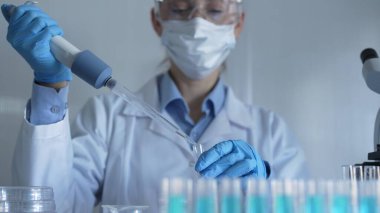 Laboratuvar önlüğü, maske, mavi eldiven ve güvenlik gözlüğü takan bilim adamı kadın test tüplerine sıvı atmak için mikropipet kullanıyor. Tıp ve Bilim.