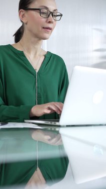 Yeşil bluz giyen iş kadını dizüstü bilgisayarda daktilo kullanıyor, vergileri ve denetim raporlarını hazırlıyor, cam ofis masasında belgeler, dikey portre görüntüsü. Vergiler, iş denetimleri.