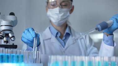Maske takan, mavi eldivenli ve güvenlik gözlüklü bir kadın bilim adamı deney tüplerine sıvı atmak için mikropipet kullanıyor. Tıp ve Bilim.