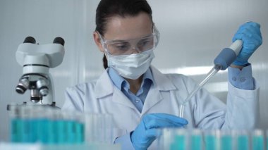 Laboratuvar önlüğü, maskesi ve güvenlik gözlüğü takan bilim adamı bir kadın deney tüpüne mavi bir sıvı atmak için mikropipet kullanıyor, portre görüntüsü. Tıp ve bilim konsepti.