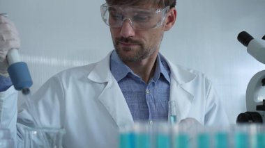 Laboratuvar önlüğü ve güvenlik gözlüğü takan bilim adamı deney tüpüne sıvı atmak için mikropipet kullanıyor. Portre görüntüsü. Tıp ve Bilim.