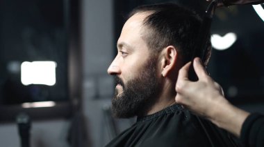 Profesyonel berber, modaya uygun bir berber dükkanında profesyonel bir makasla, sakallı bir adam için modern bir saç stili hazırlayan bir müşterinin saçını ustaca kesiyor.