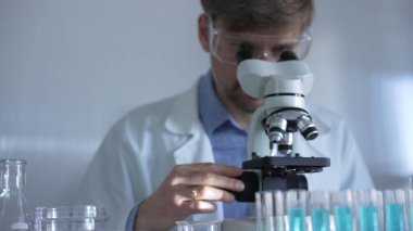 Laboratuvar önlüğü, maskesi ve güvenlik gözlüğü takan bir bilim adamı laboratuvar ortamında test tüpleriyle çevrili bir mikroskop kullanıyor. Tıp ve bilim konsepti.