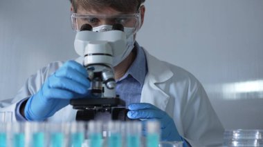 Laboratuvar önlüğü, maske, mavi eldiven ve güvenlik gözlüğü takan bilim adamı laboratuarda test tüpleriyle çevrili bir dürbün kullanıyor. Portre görüntüsü. Tıp ve bilim konsepti.