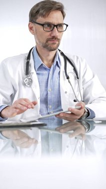 Beyaz önlük, gözlük ve steteskop altında mavi gömlekli doktor modern sağlık ortamında dijital tablet kullanıyor. Hasta verileriyle, dikey portre görüntüsüyle etkileşimde. Tıp ve Bilim.