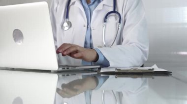 Doktor kadın, doktor önlüğünün altına mavi bir bluz giyiyor, sağlık hizmetlerinde teknolojiyi kullanıyor, elleri bilgisayarında stetoskop, gözlük ve pano ile daktilo kullanıyor. Tıp konsepti.