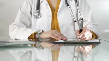 Beyaz bir sağlık önlüğünün altına sarı bluz giyen kadın doktor, bir pano kullanıyor ve yakın plandaki cam masaya notlar alıyor. Tıp konsepti