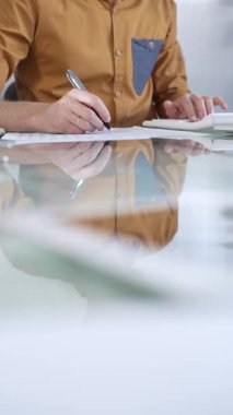 İş adamı muhasebeci gündelik sarı gömlek giyiyor ofis cam masasındaki hesap makinesini kullanarak mali raporları dikkatlice inceliyor, dikey yakın çekim görüntüsü. Vergiler, iş denetimleri.