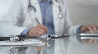 Stetoskop ve ilaç geçmişi kayıtları cam bir masanın üzerinde duruyor. Klinik ofis arka planında dizüstü bilgisayarla çalışan bir erkek doktorun önünde. Tıp konsepti.