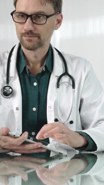 Modern sağlık ortamında dijital tablet kullanan yeşil önlüklü bir doktor hasta verileriyle, dikey portrelerle etkileşime geçiyor. Tıp konsepti.